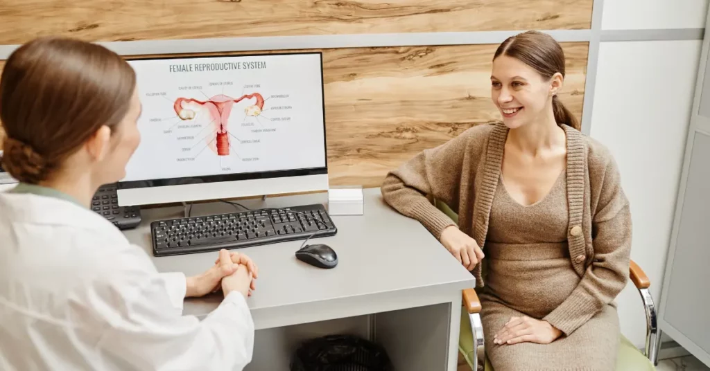 Female fertility exam consultation with a pregnant woman speaking to a doctor beside a reproductive anatomy diagram.