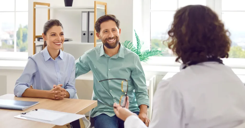 Low stim fertility treatment couple smiling during a relaxed fertility consultation with a doctor in a bright office.